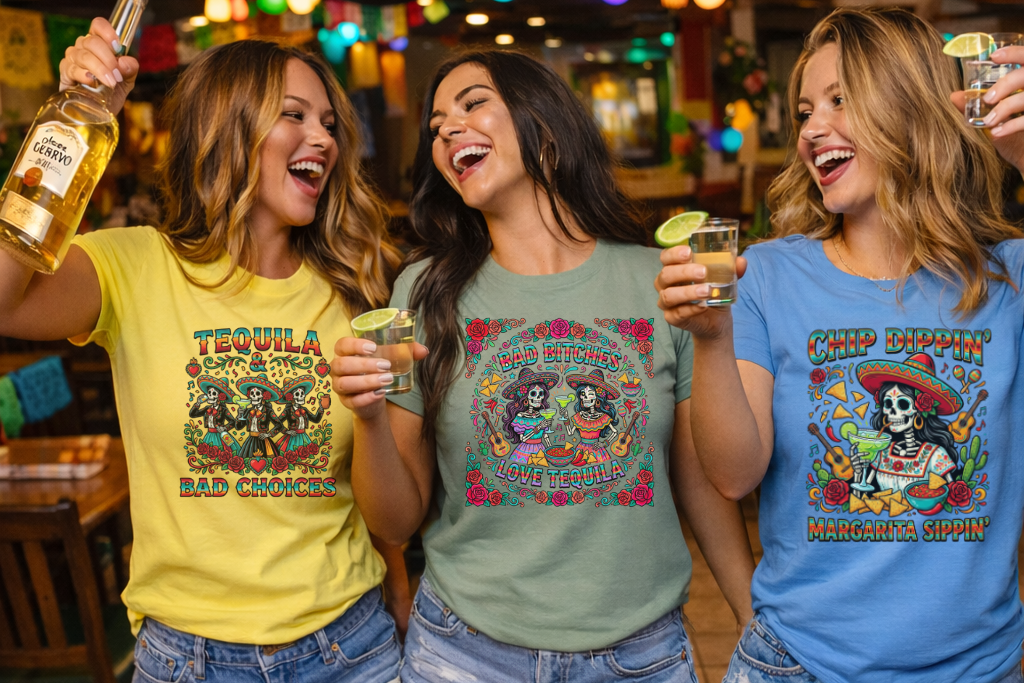 Three women in colorful t-shirts with tequila-themed designs, holding drinks in a festive bar setting.